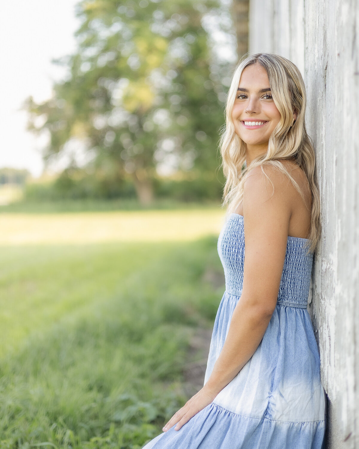 high school senior female, standing against a gray and white barn, with long blonde hair, wearing a white and blue sleeveless dress, smiling at the camera