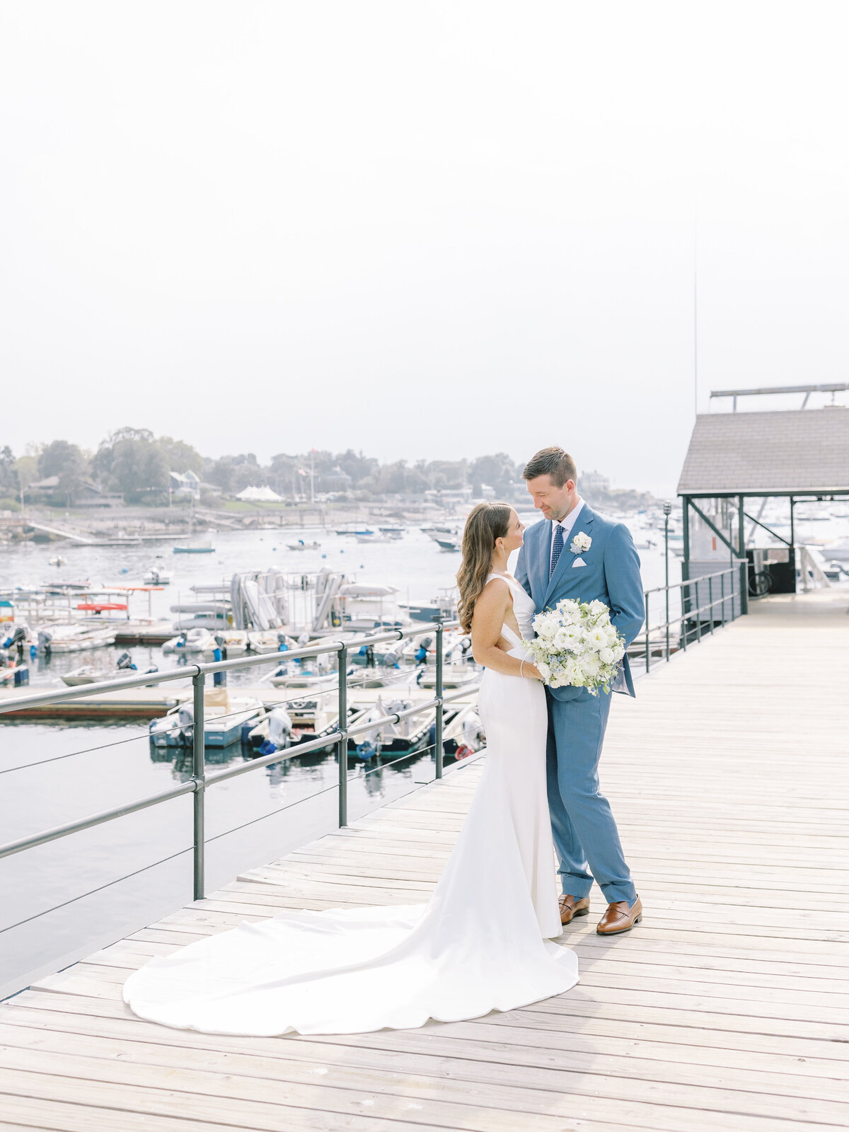 Bride and groom standing together and smiling at each other on a dock in front of boats on the water