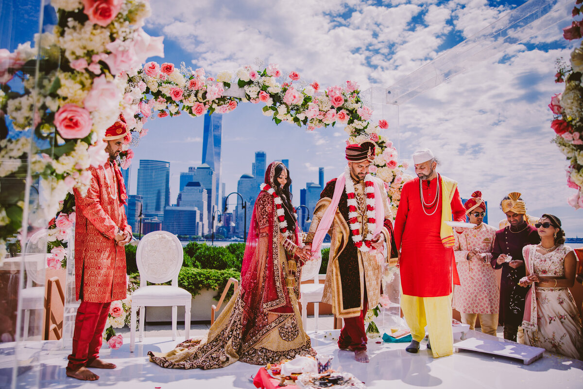 Indian bride and groom walking around the fire (pheras) during their hindu wedding ceremony under a floral glass mandap with the nyc skyline in the background at jersey city hyatt