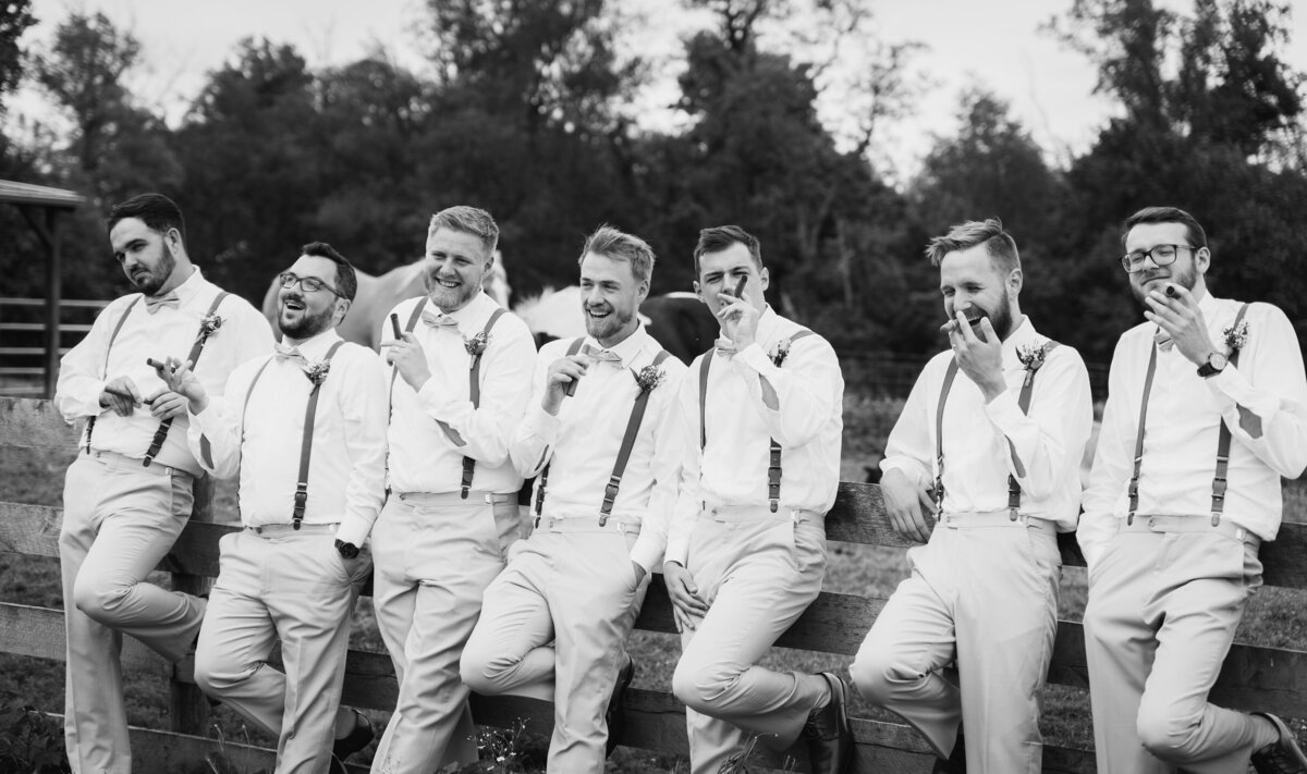 The groom, Alex Cox, and his groomsmen lined up against a fence with cigars at Alex's wedding at Peacock Ridge in Lawrence, Ohio.