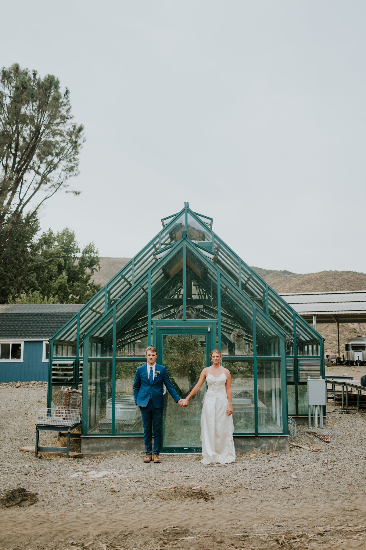 Bride and groom stand apart while holding hands in front of greenhouse.