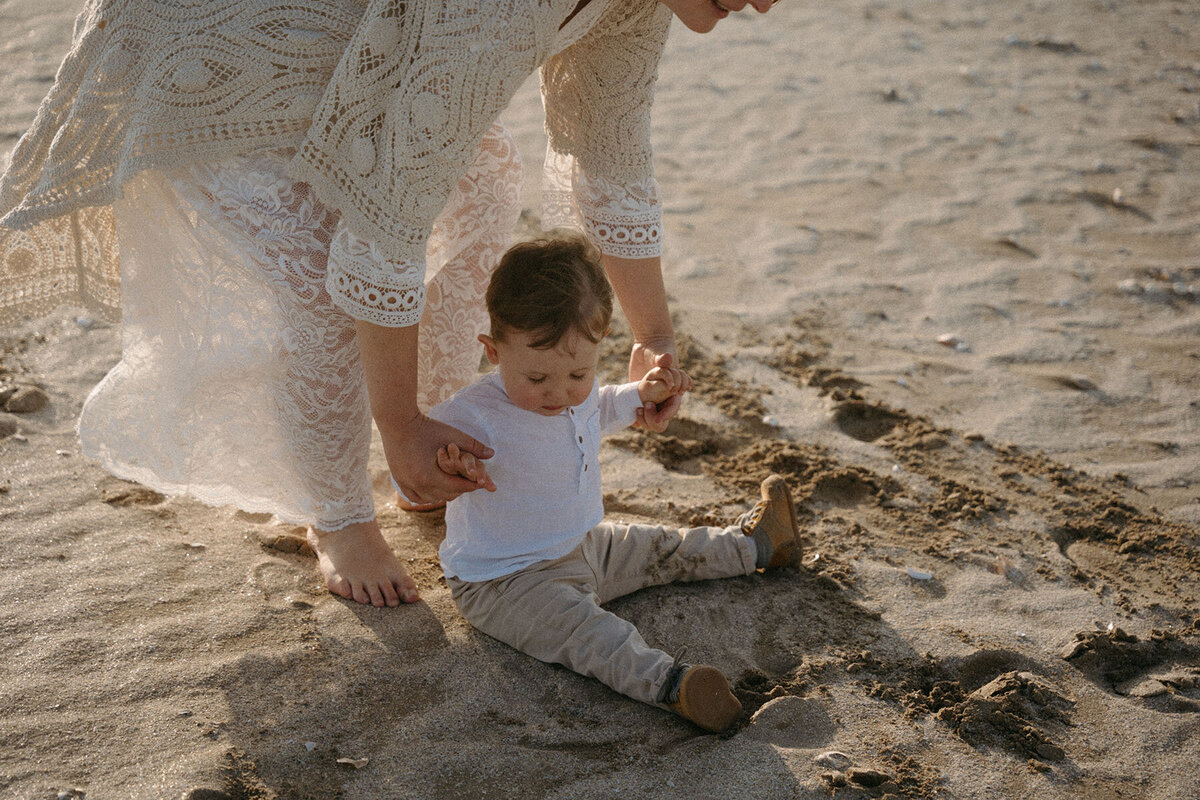Petit garçon assis dans le sable retenu par les bras de sa maman. Séance photo famille à la mer. 