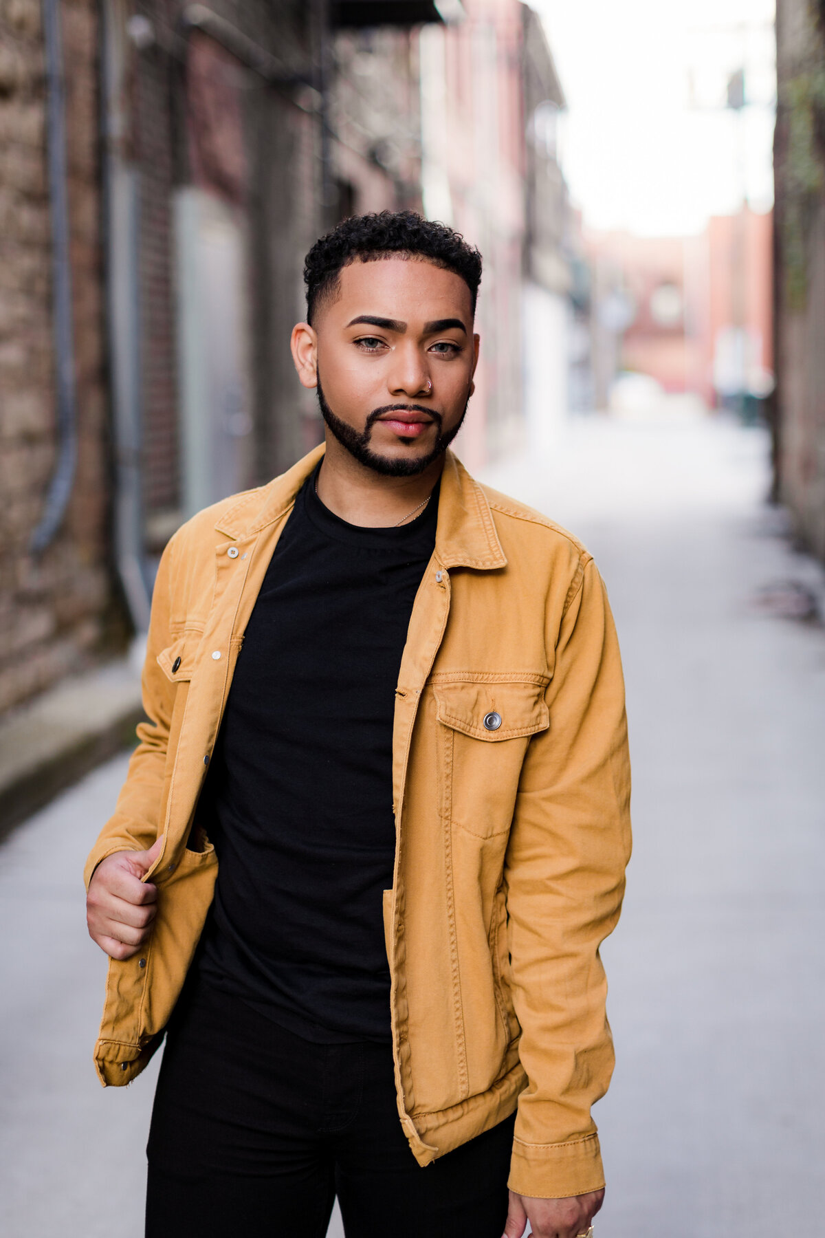 Tav poses for the camera with a serious expression in an alley while holding his yellow jacket with one hand.