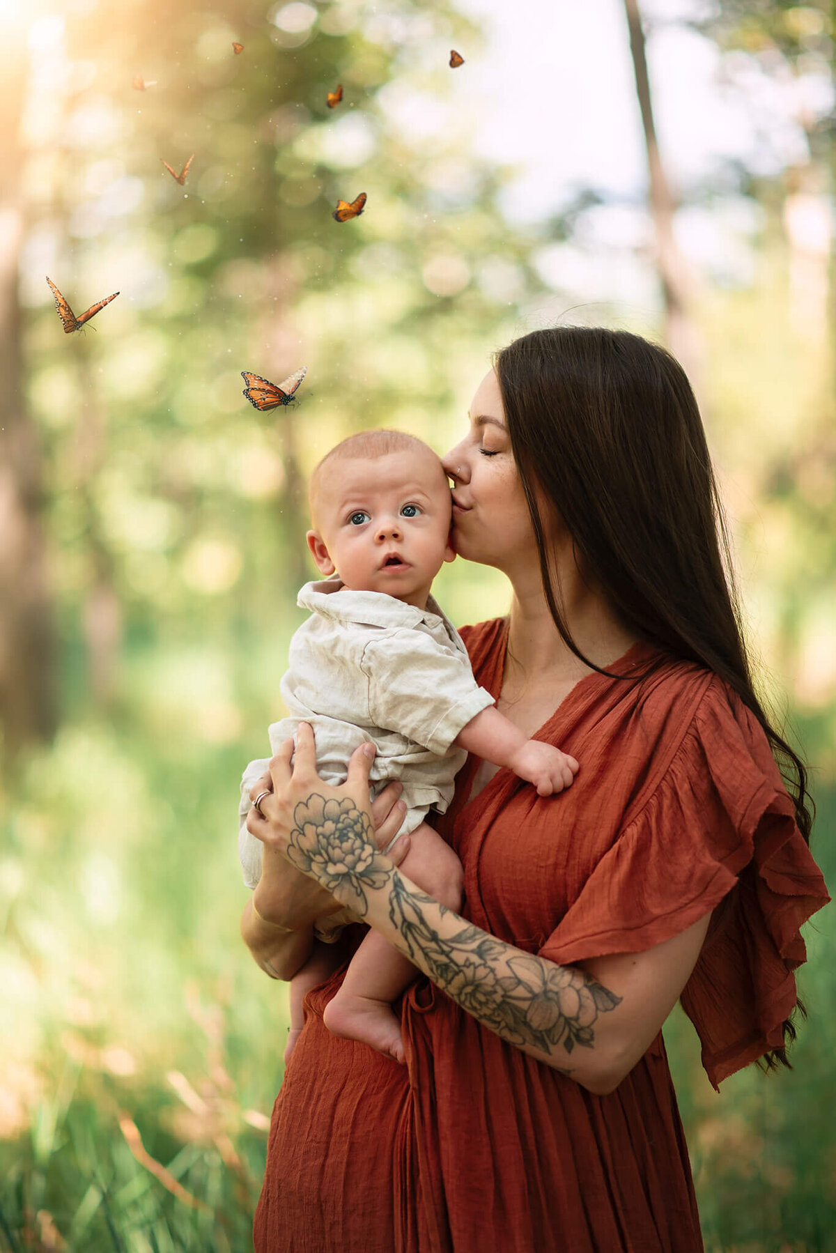 A beautiful springtime portrait of a mother and son captured in kamloops by Brittany Danielle Photography. Mom wears a flowy rust coloured dress whule holding her infant son dressed in linen. She kisses his cheek while he looks off at a group of butterflies