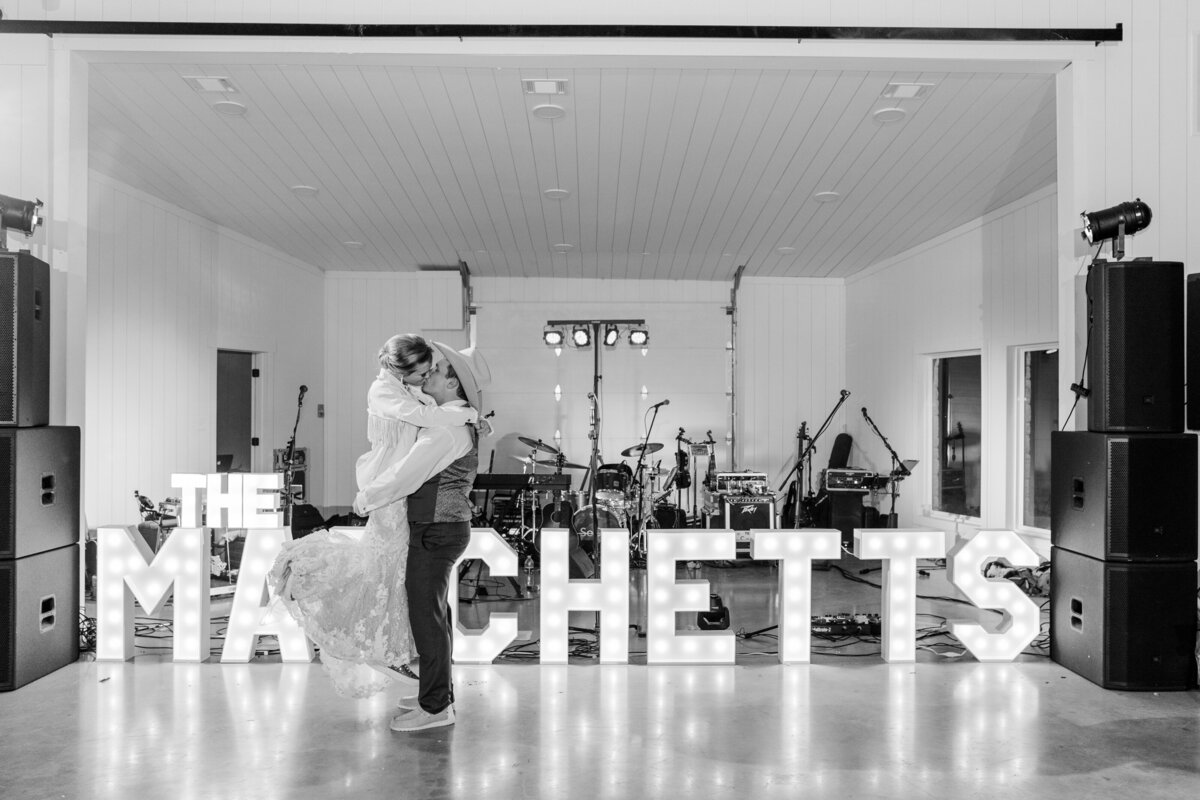 Black and white photo of Groom picking up Bride in front of marquee letters at their reception at The Ranch House