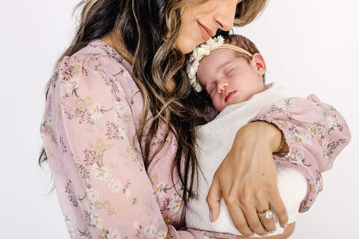 A woman in a floral dress gently cradles a sleeping baby wrapped in a white blanket. The baby wears a floral headband. They are against a plain white background.