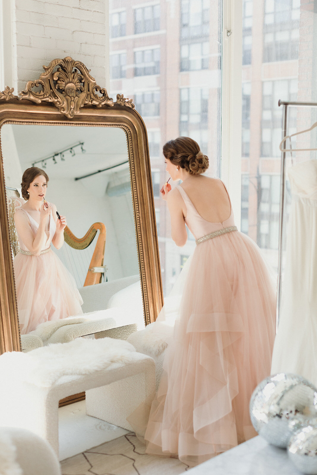 light skinned woman in pale pink gown is leaning toward a large gilded mirror and applying lip gloss. Her harp can be seen in the reflection in the mirror.