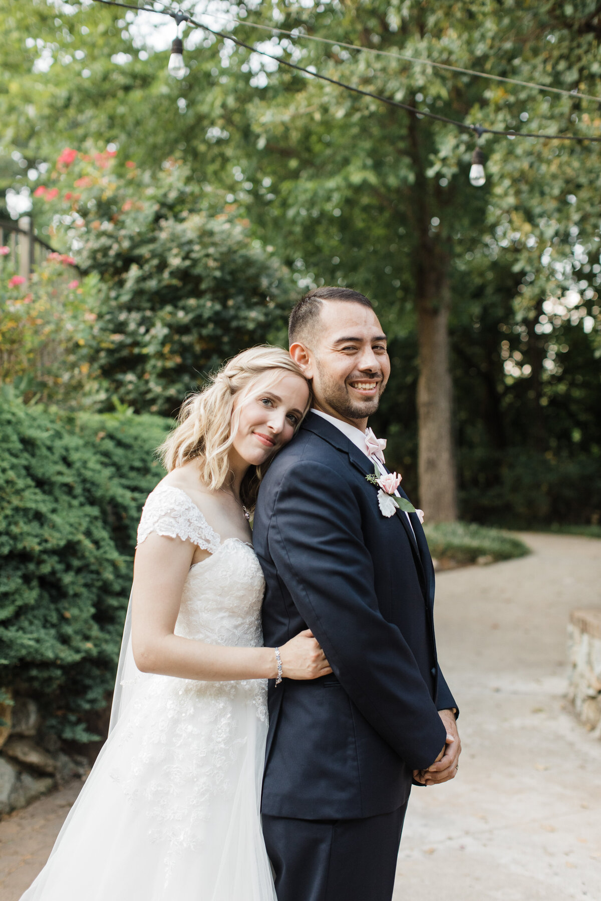 Portrait of a bride and groom on their wedding day in Fort Worth, Texas. The groom is smiling largely and standing tall while the bride is smiling coyly and leaning her head on his shoulder. The bride is wearing an intricate, white dress while the groom is wearing a dark suit with a boutonniere.