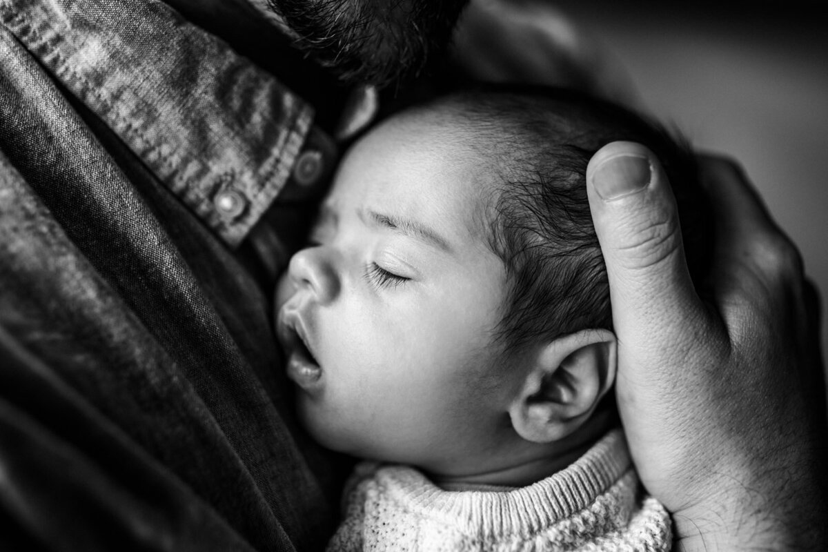 newborn sleeping with mom slighting open on dad's chest with dad's hand wrapped around baby's head
