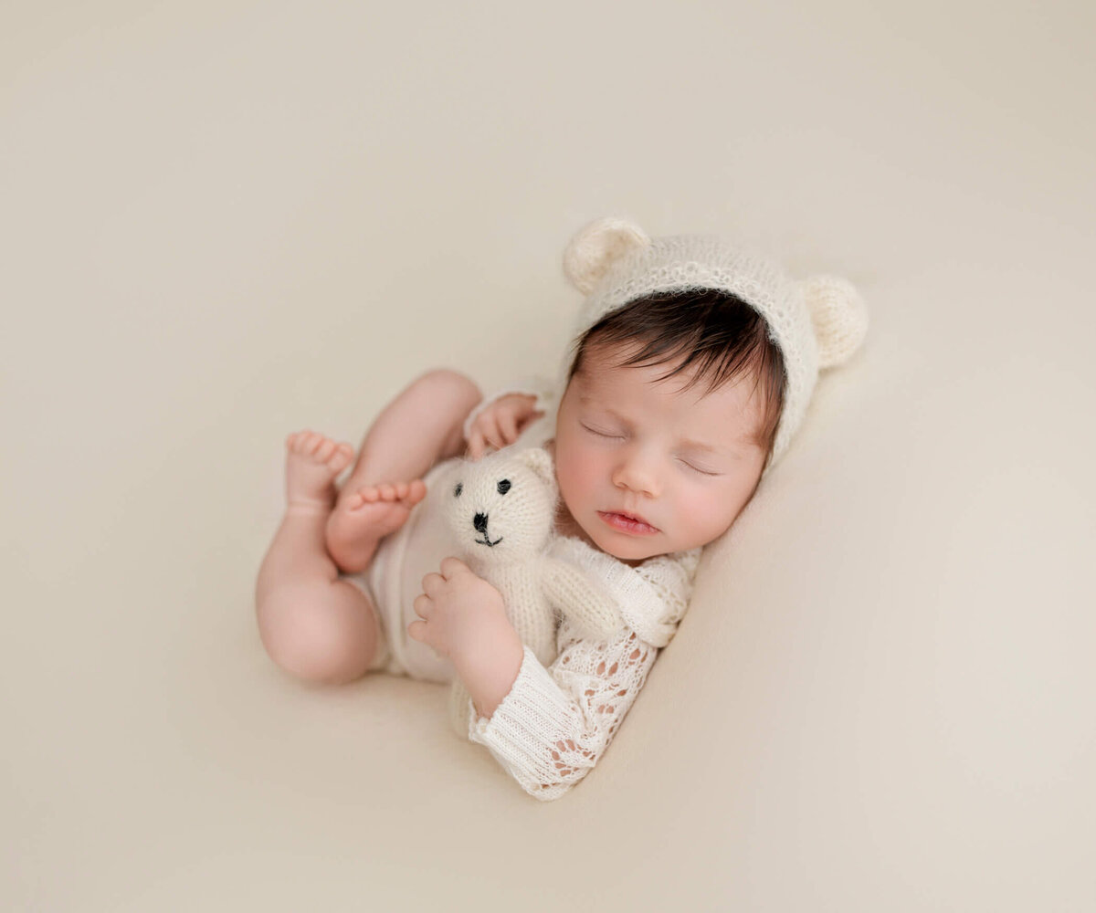 A sleeping newborn baby in a white onesie and bear ears hat, cuddling a small white teddy bear. The baby is peacefully resting on a soft, beige background.