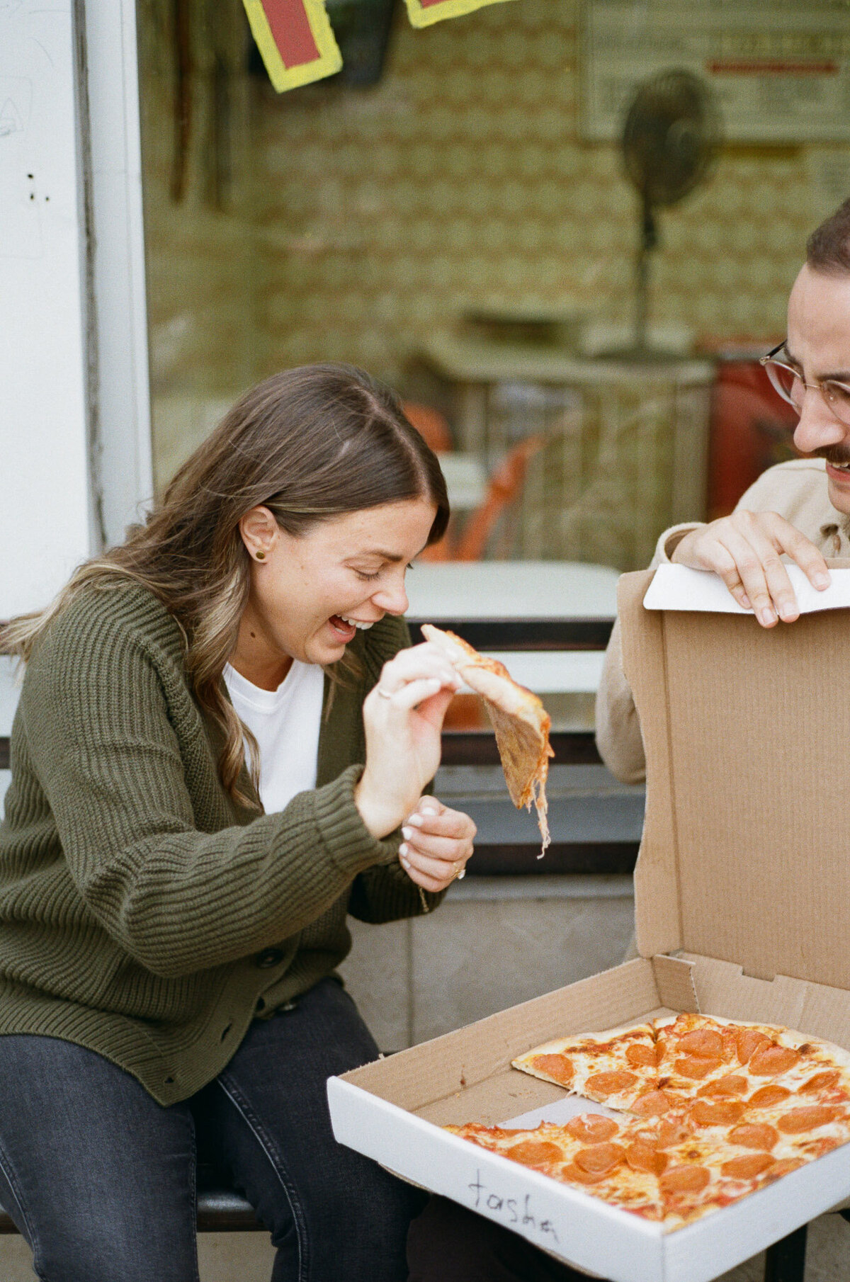 Two people sit outside, enjoying slices of pepperoni pizza from an open box to celebrate their engagement in Trinity Bellwoods, captured by Toronto engagement photographer, Jennifer van Son.