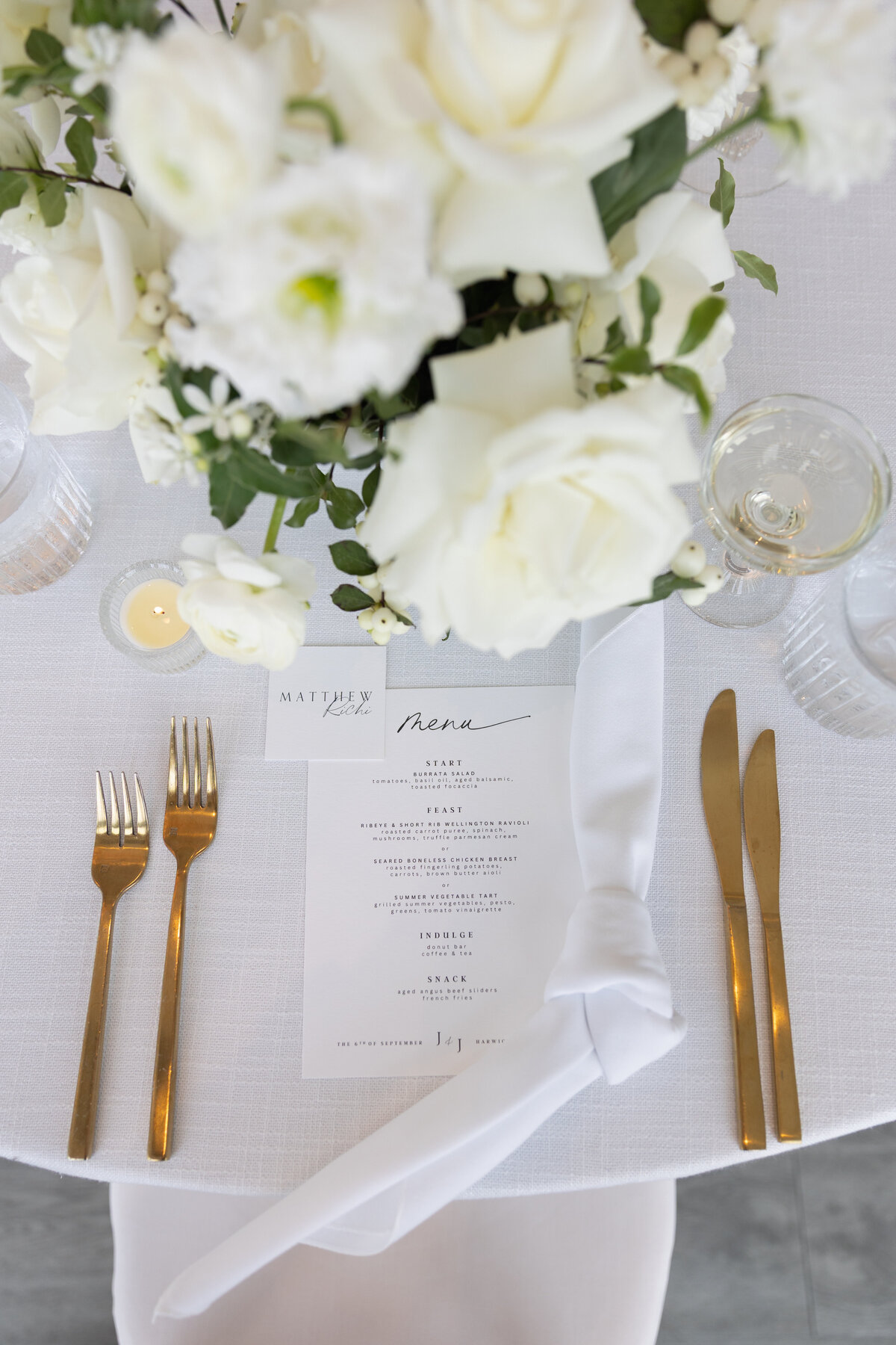 Rhode Island Wedding Photographers | Kelsey Sheehan Photography Timeless Rhode Island Weddings | Elegant wedding table setting with white roses, a neatly tied napkin, a gold cutlery set, and a printed menu on a white cloth. Sophisticated and serene ambiance.