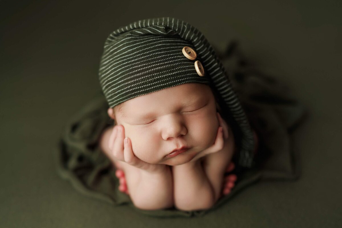 Squishy newborn boy with a green sleepy cap posing for his photo session