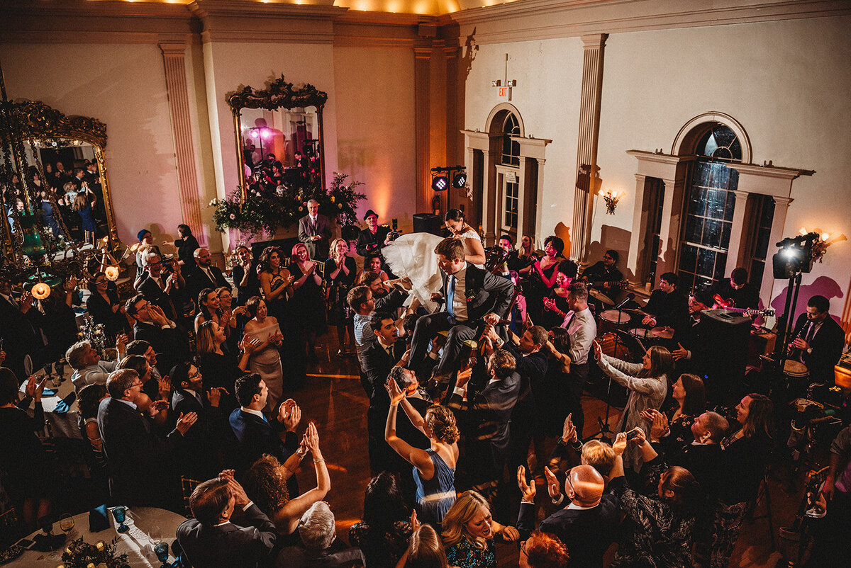 bride and groom are lifted up on chairs during their wedding reception at hamilton hall photo by cait fletcher photography