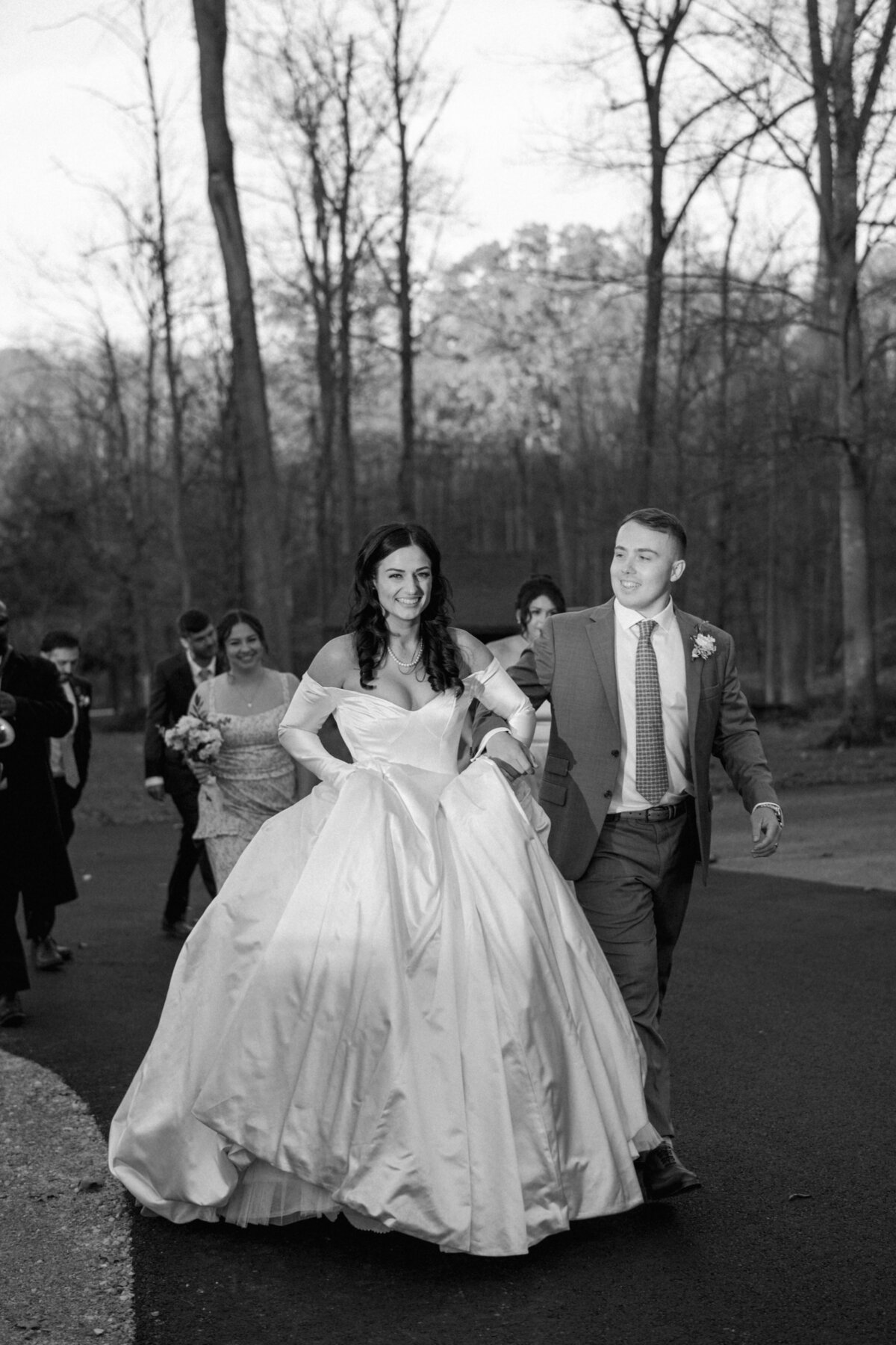 Black and white photo of a bride in a flowing gown and groom walking joyfully outdoors, accompanied by smiling guests in a wooded setting.