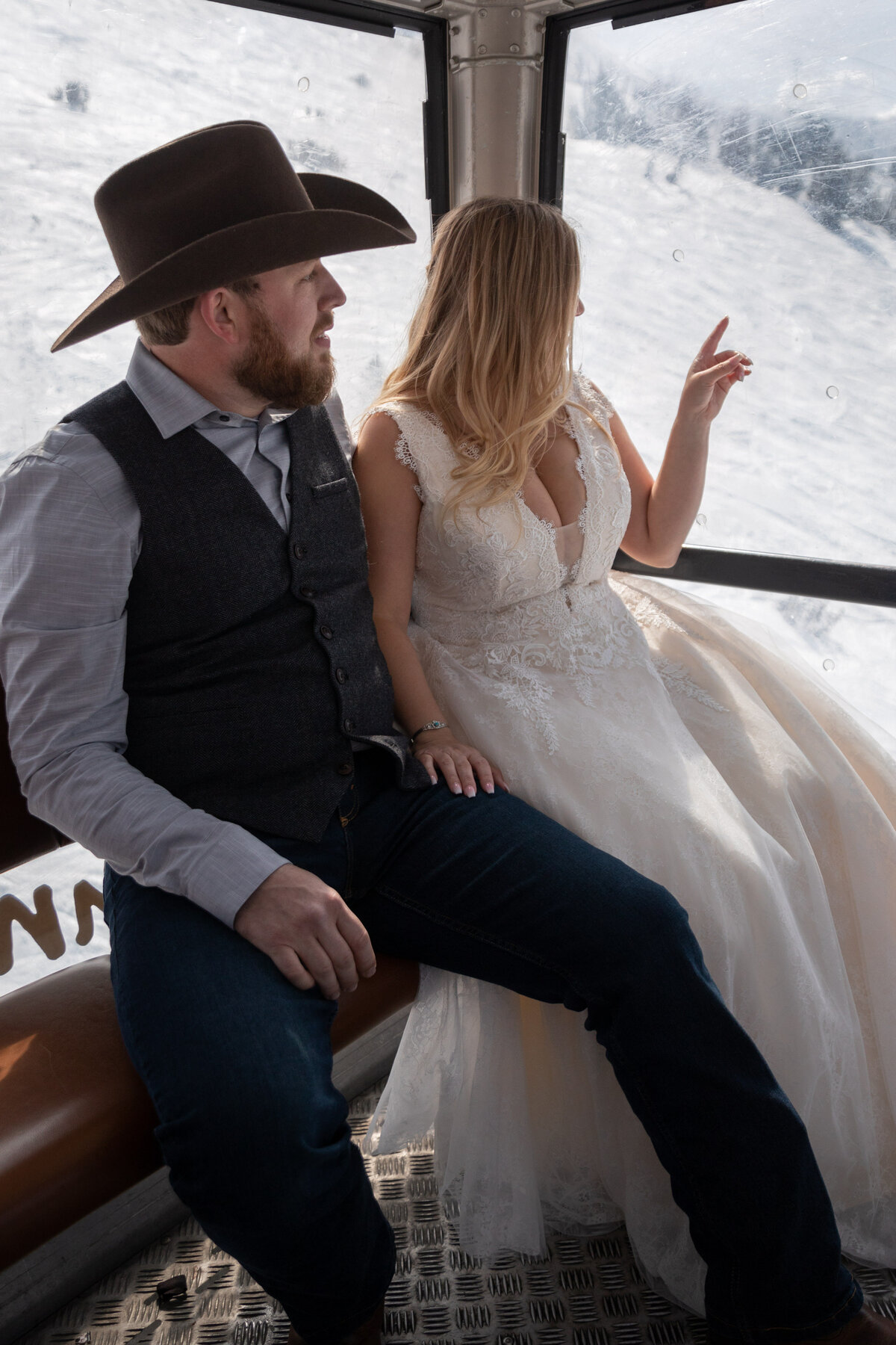 A bride and groom sit next to each other on a gondola traveling down a mountain while the bride points out the window.