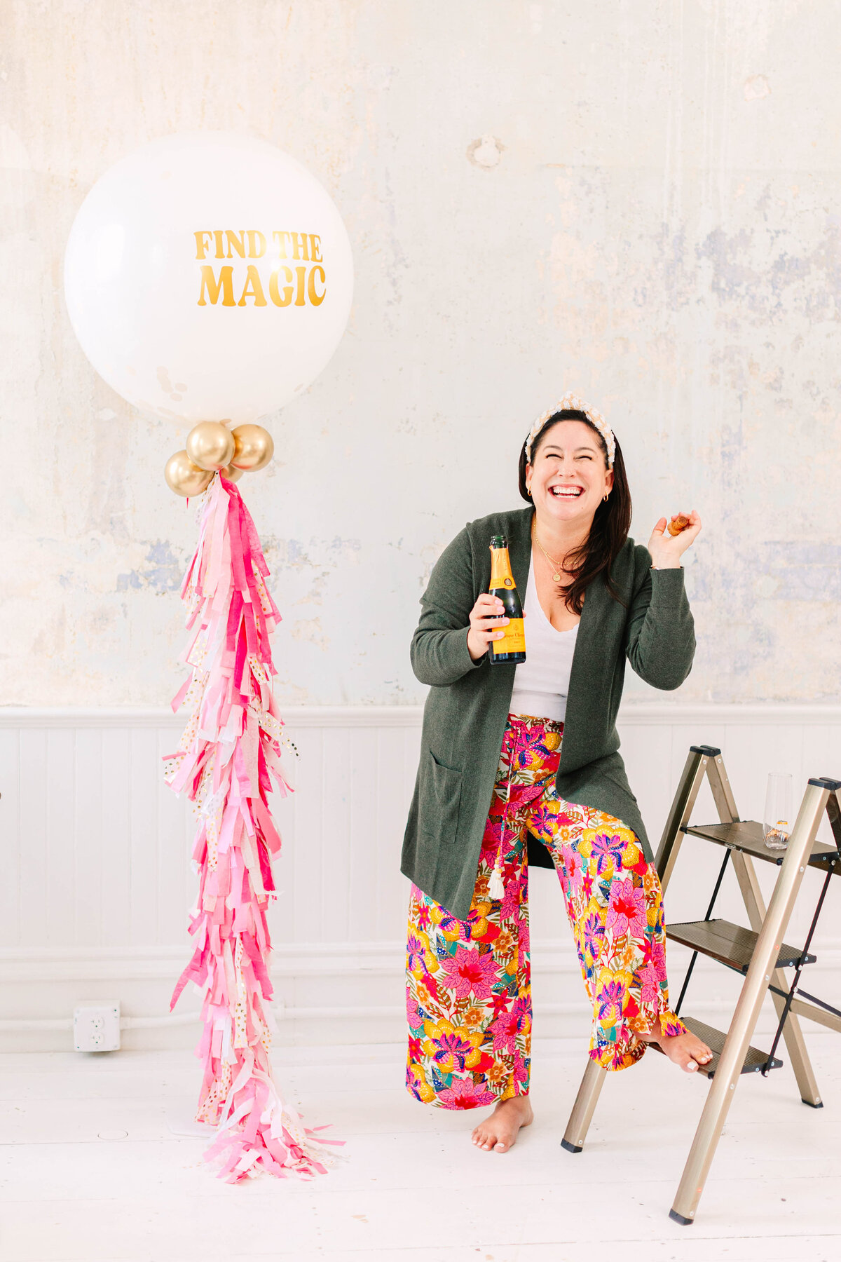 A woman in colorful floral pants and a green cardigan sits on a step ladder, holding a bottle of champagne and laughing. A large white balloon with "Find the Magic" in gold lettering and pink tassels stands beside her against a textured pastel wall.