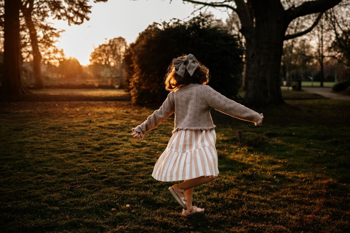 Backlit image of a little girl with curly hair and a big bow, joyfully spinning in a park as the sun sets behind the tree