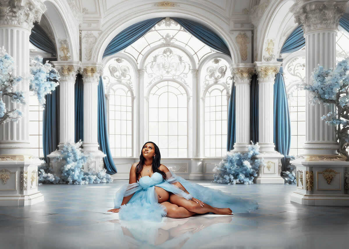 Pregnant woman in a baby blue tulle gown sitting down in a white and blue decorated room with pillars and large windows