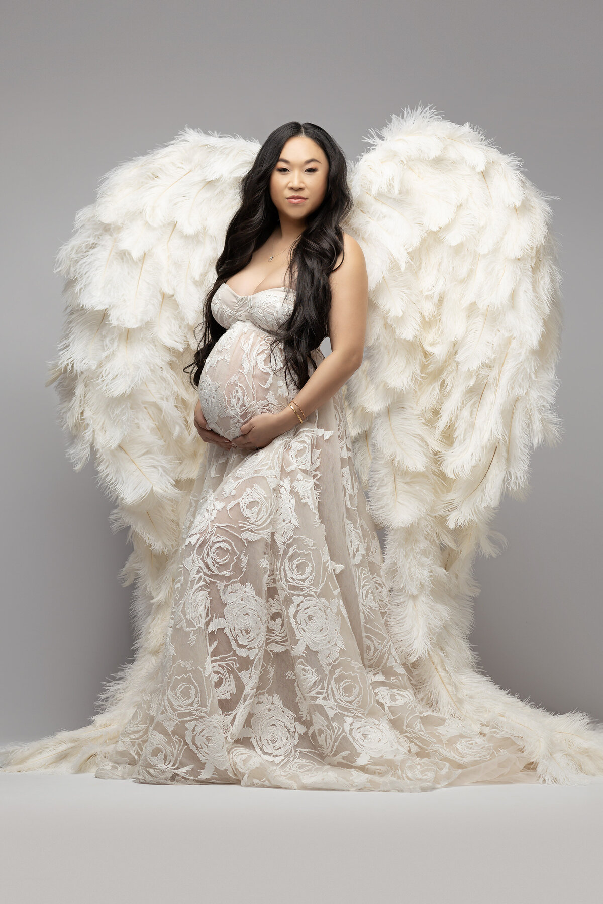 A beautiful woman in a lace gown stands with large, white angel wings against a gray background, conveying a serene and ethereal mood. Dallas TX maternity photography by Lindsay Walden.