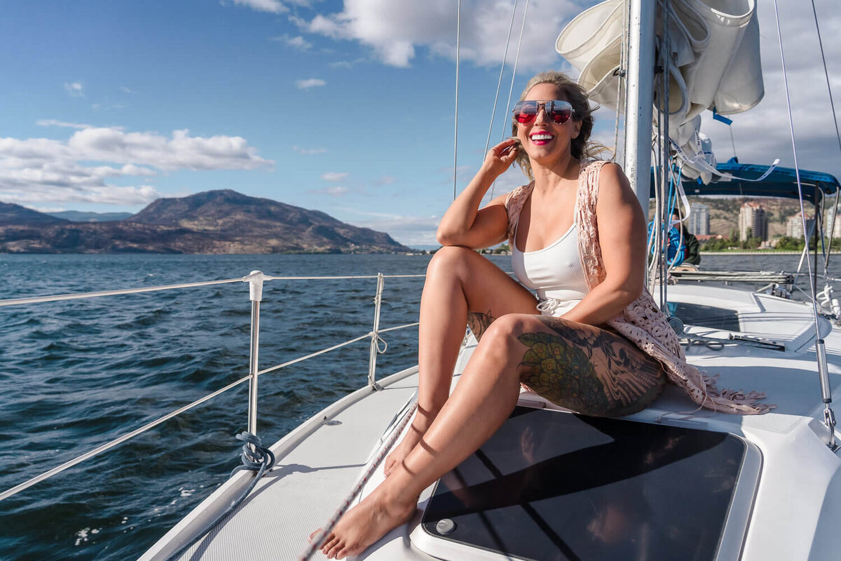 Happy woman in white tank sitting on sailboat deck enjoying sunshine on Okanagan Lake.