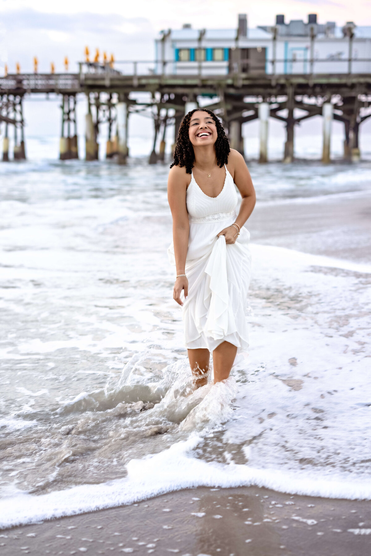 High school senior laughing and holding her dress near Cocoa Beach Pier, eyes lifted, carefree joy and glowing sunshine.