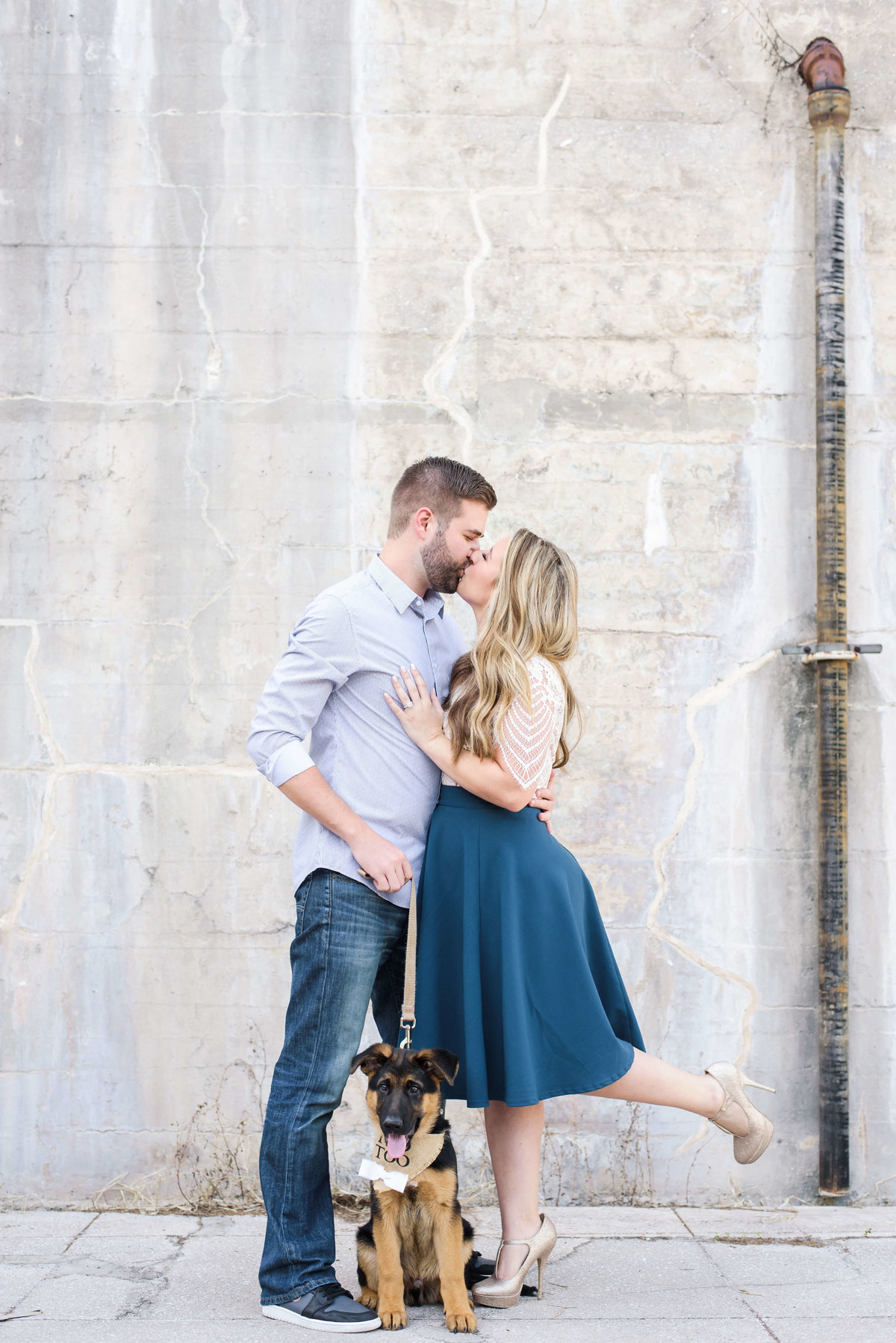 Woman lifts her leg while she kisses her fiance as their puppy looks on during this super sweet engagement session in Sarasota