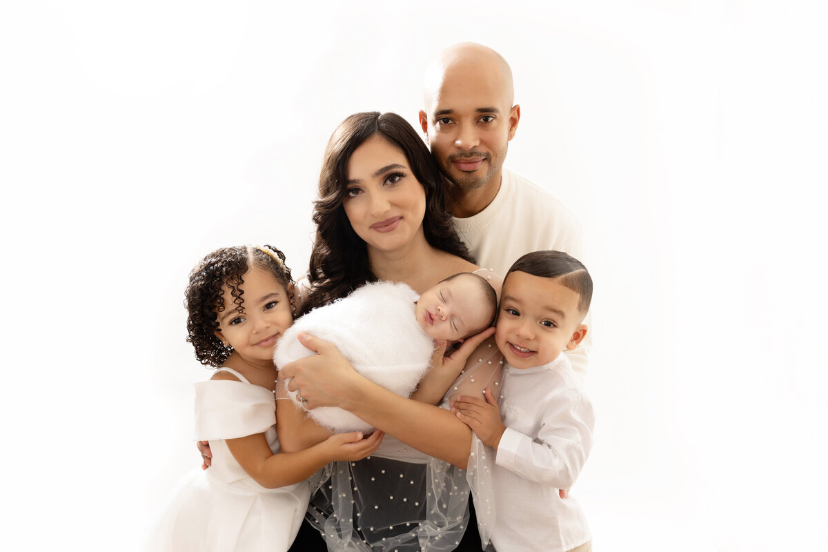A mom and dad stand in a studio with their newborn baby and toddler son and daughter