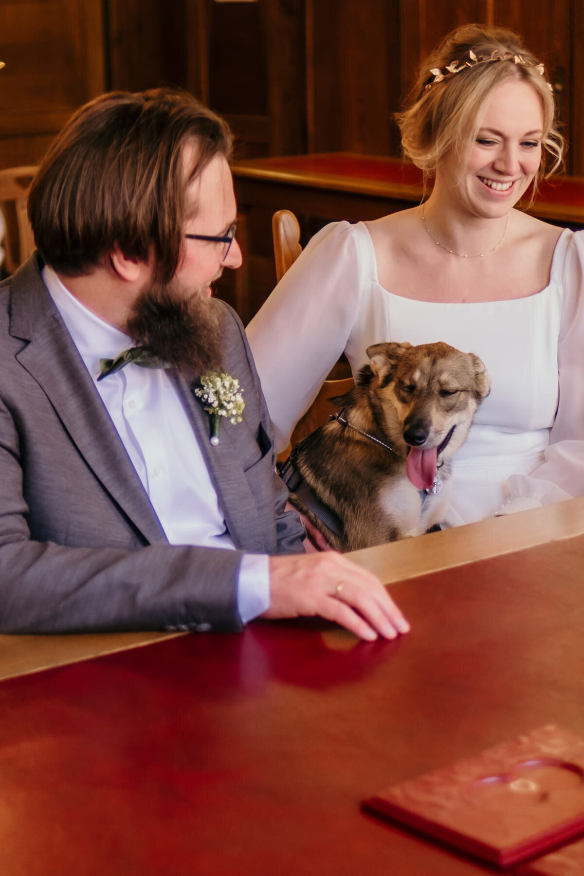 Am Trautisch des Standesamts sitzt das lächelnde Hochzeitspaar mit ihrem Hund in der Mitte.