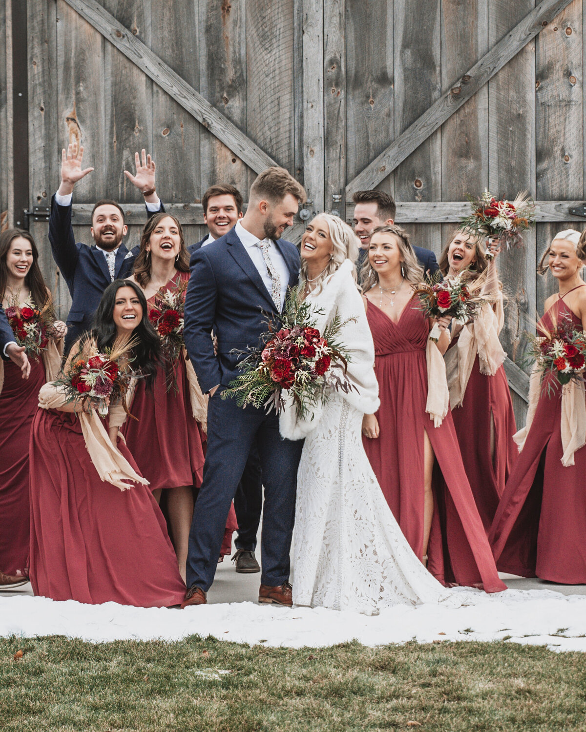 A joyful winter wedding party celebrating, with the bride and groom sharing a sweet kiss as their friends cheer around them, against a rustic wooden barn backdrop taken by jen Jarmuzek photography a Minneapolis wedding photographer