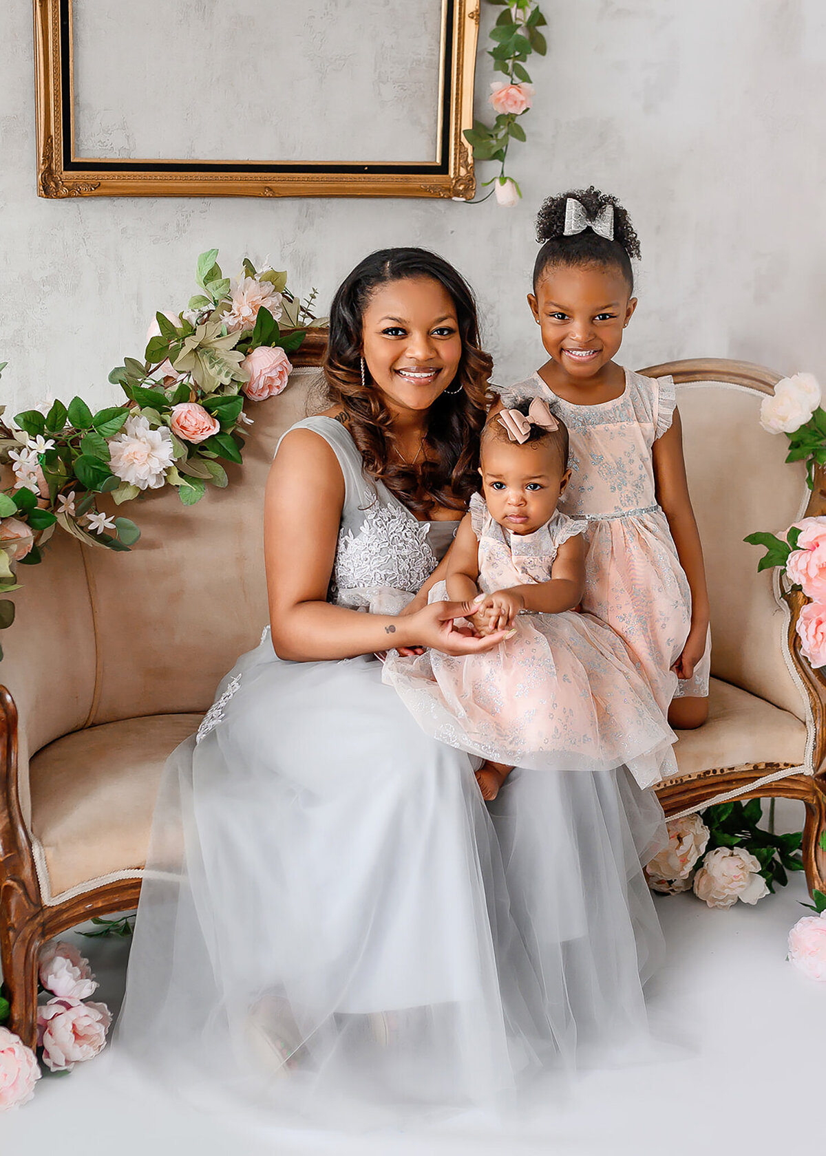lovely portrait of a mom sitting on a vintage couch with floral garland holding her younger daughter and older daughter is kneeling next to her