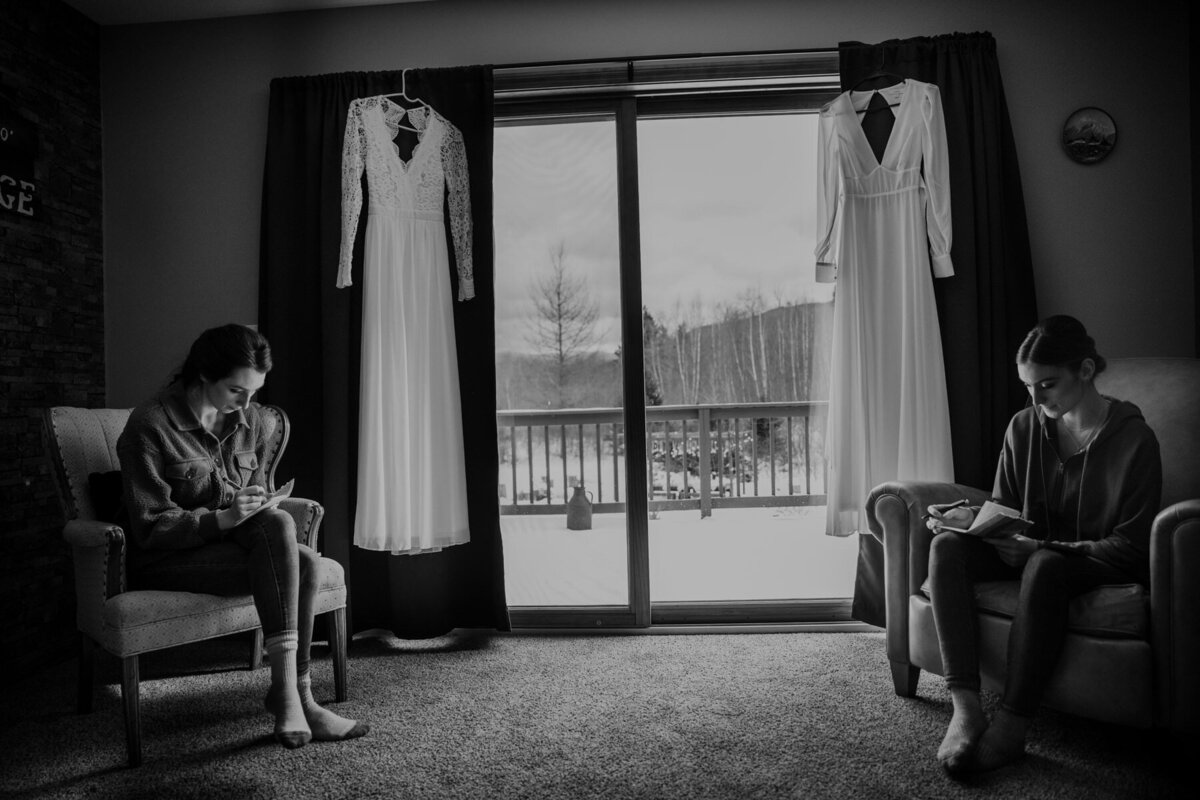 Two brides sit in the same room writing their vows for their elopement day. Their dresses are hanging in the room with them.
