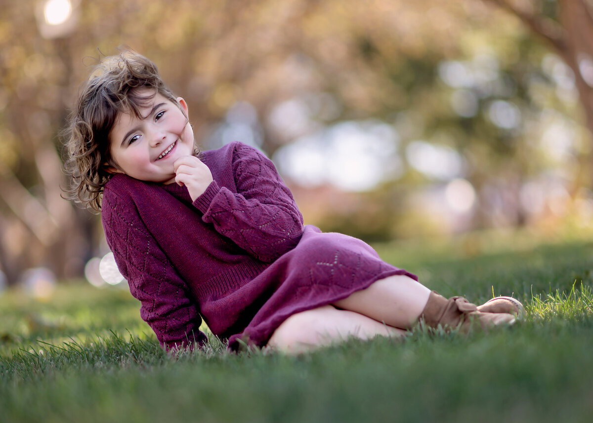 Girl in a purple outfit with her hand under her chin sitting on grass with a blurred background.