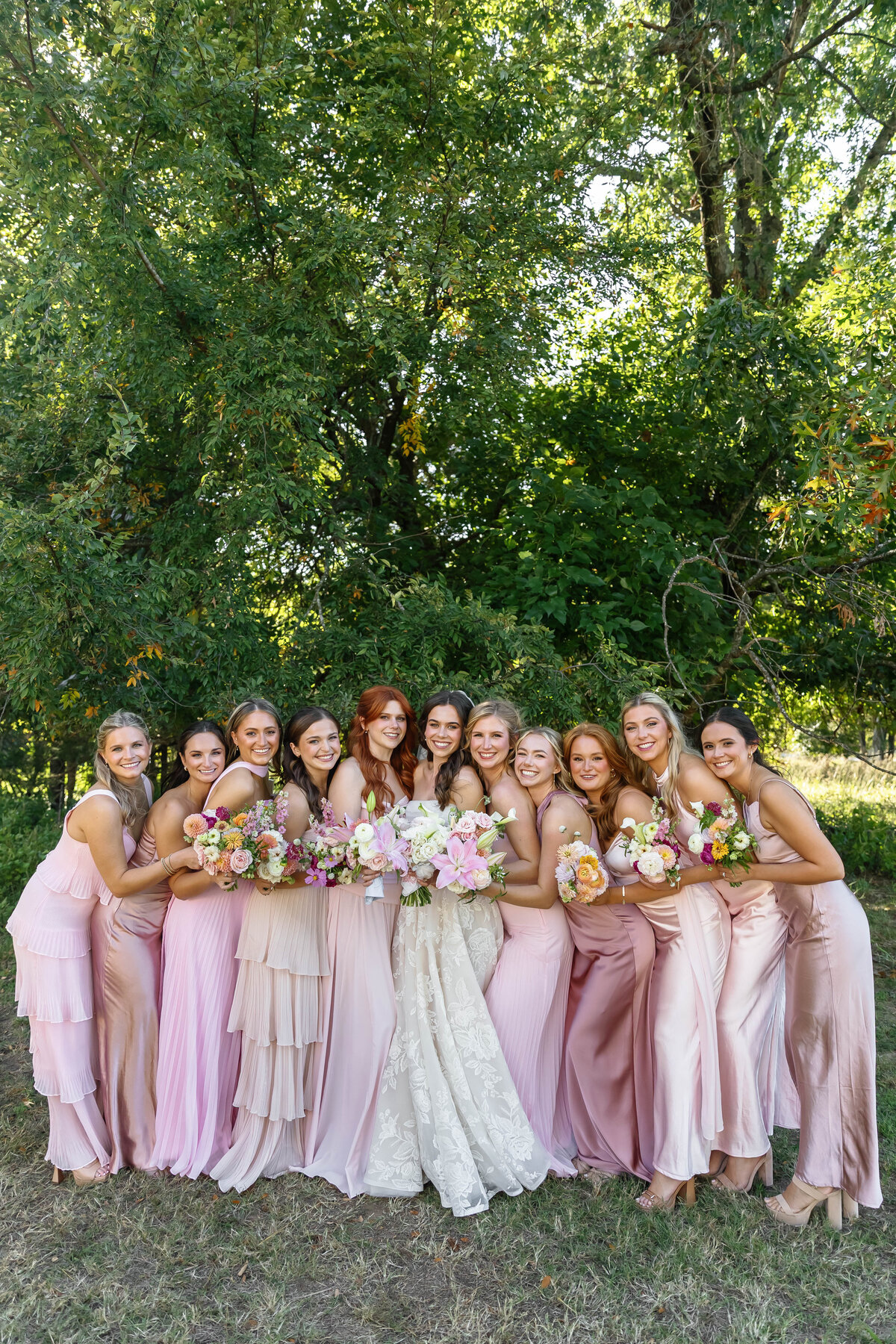 Full bridal party of bridesmaids surrounding the bride, all wearing mix-and-match pink dresses, holding fresh floral bouquets during outdoor wedding portraits under lush green trees.
