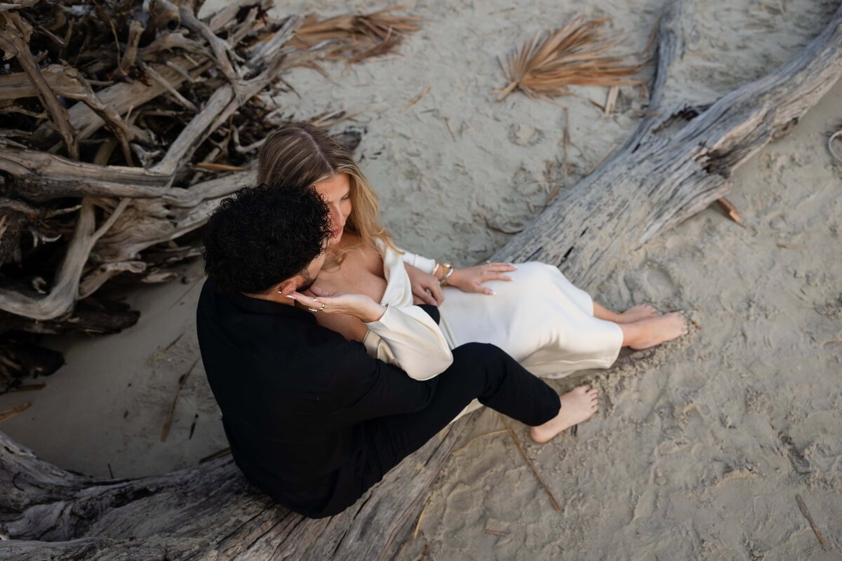 An engaged couple shares a quiet, intimate moment while sitting barefoot on a large piece of driftwood at Driftwood Beach on Jekyll Island. The bride-to-be, in a silky off-the-shoulder white dress, leans into her fiancé’s embrace as he gently holds her face. Surrounded by sand and weathered palm fronds, the earthy tones and relaxed pose create a dreamy, coastal atmosphere. Captured by Heartful Films, a Jacksonville engagement photographer and videographer known for natural, elegant storytelling in scenic locations.