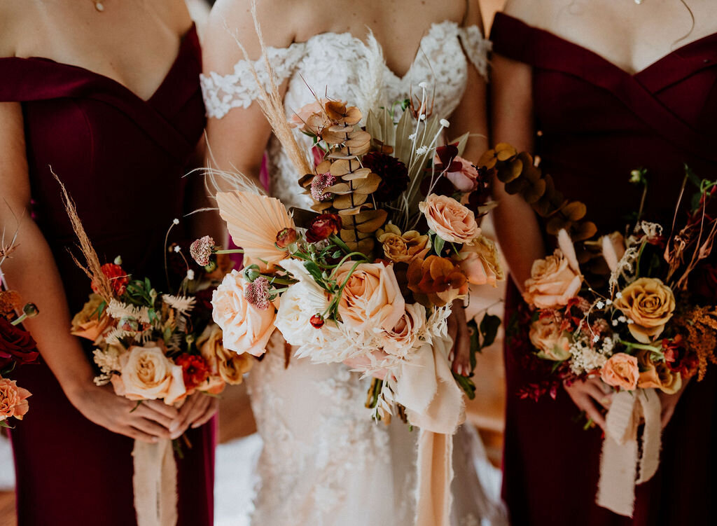 A bride and two bridesmaids wearing burgundy dresses carry terracotta and peach fresh and dried flower bouquets at a barn wedding at Stonefields Barn in Ottawa Ontario