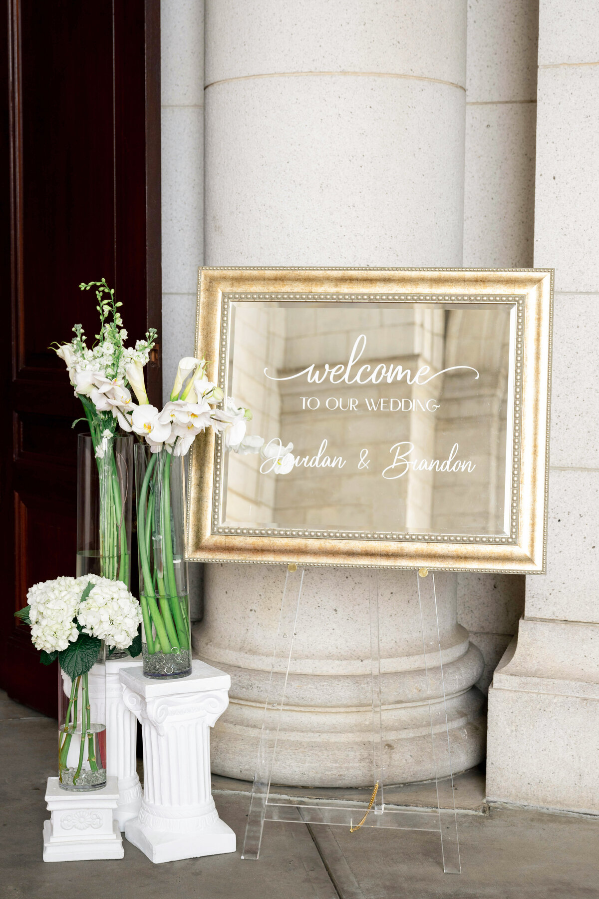 A large, elegant mirror with a gold frame displays the words Welcome to Our Wedding alongside floral arrangements of white lilies and hydrangeas on white pedestals, set against a columned building entrance.