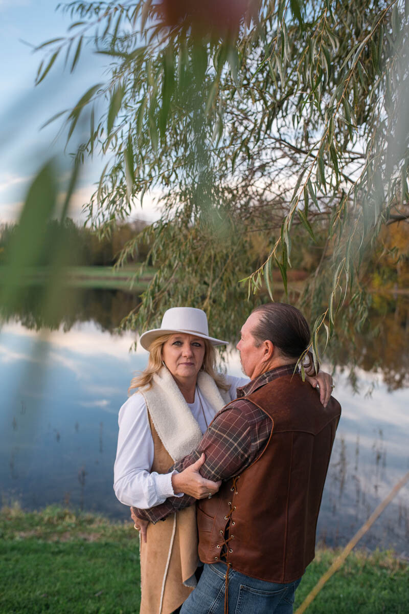 Couple holds one another closely by a lake under willow tree branches