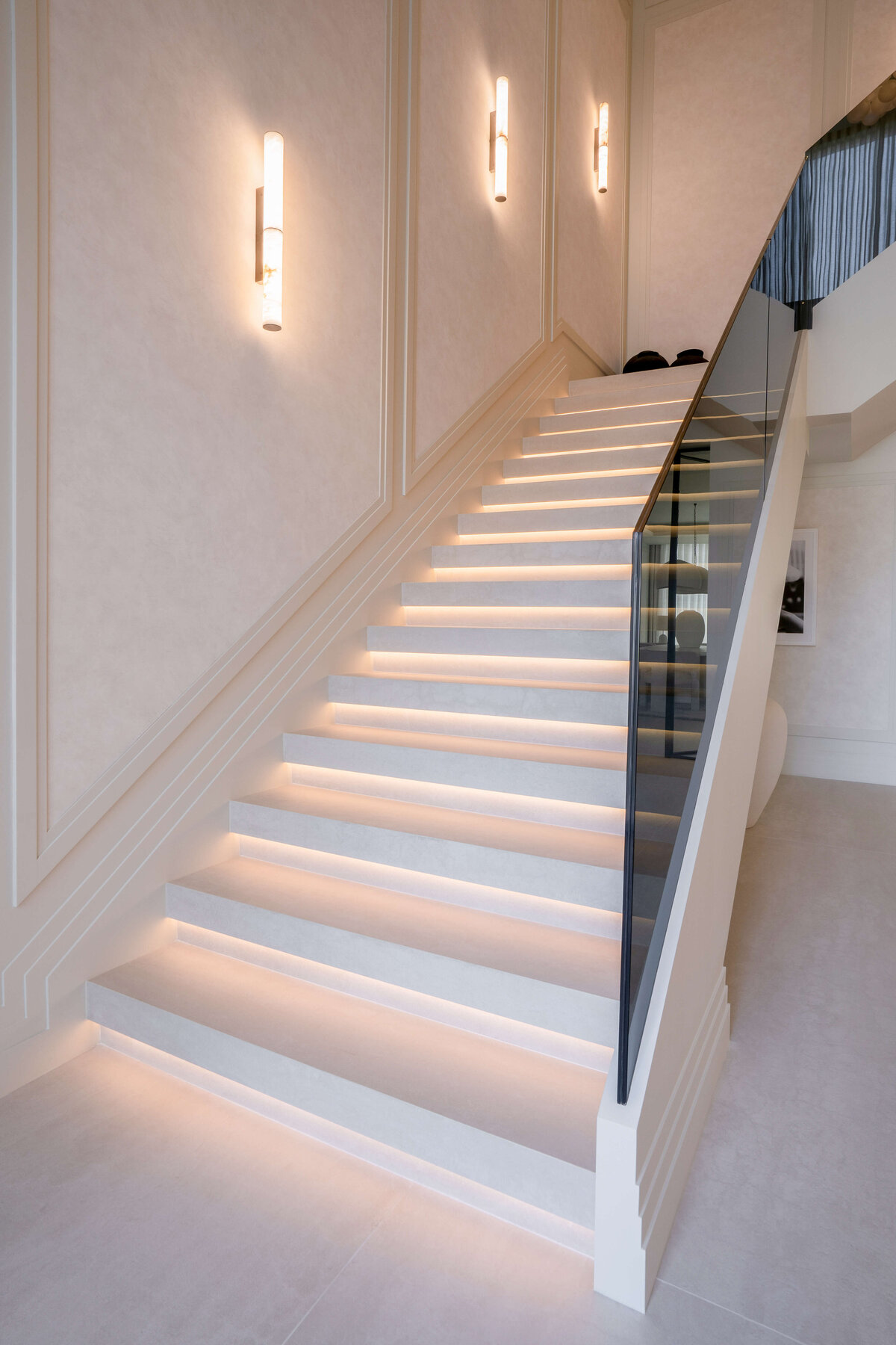 A modern staircase with a minimalist design featuring wide, white steps illuminated by soft, integrated lighting. The stairs are bordered by a sleek, black glass railing on the right side. The walls are adorned with vertical, cylindrical wall sconces that emit a warm glow.