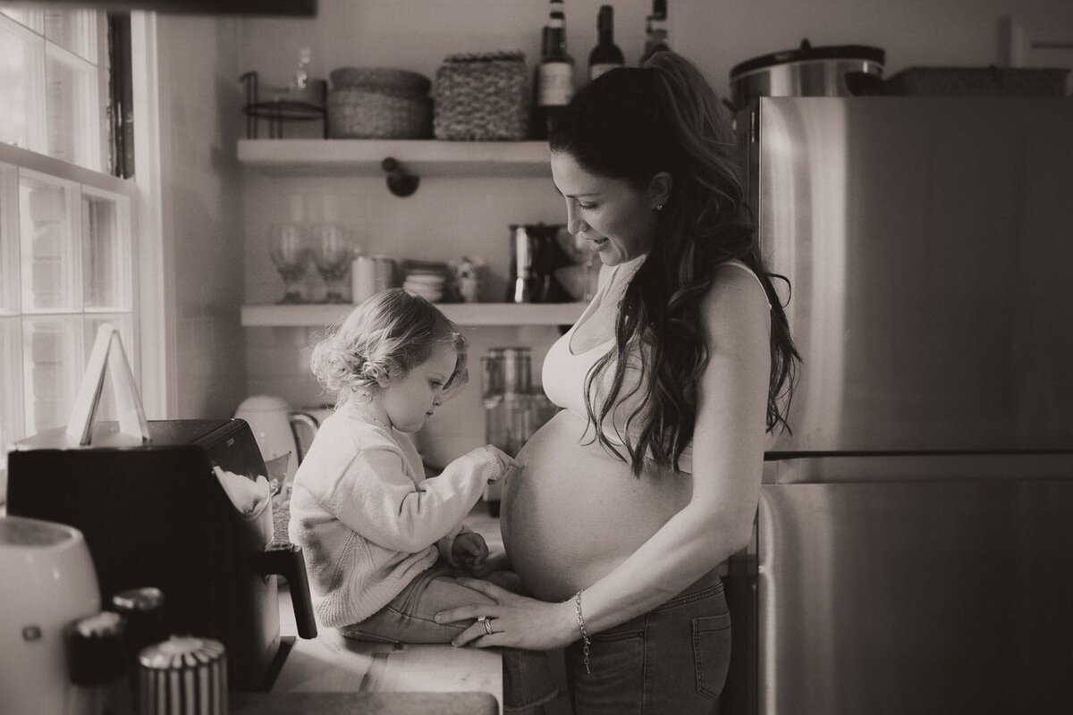 A pregnant woman smiles at a child while standing in a kitchen. The child is sitting on the counter, touching her belly. The room features shelves with various kitchen items.