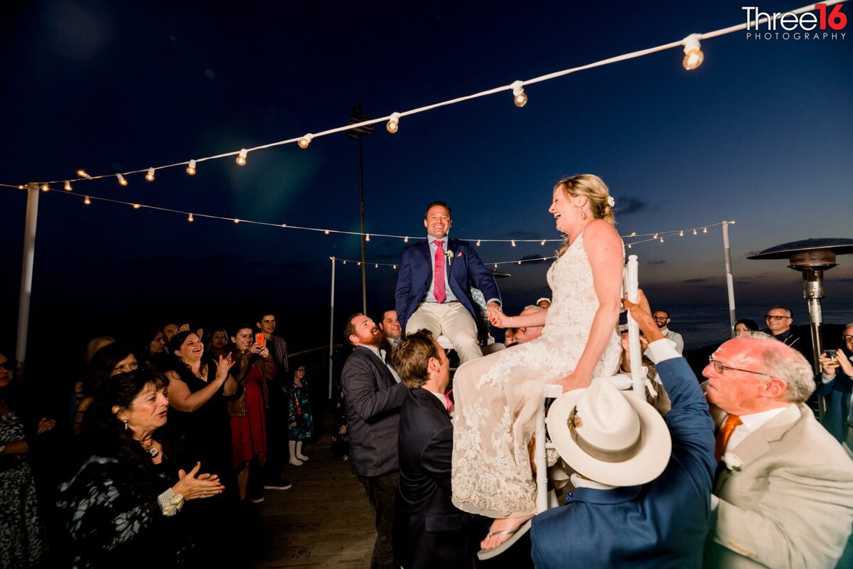 Both the Bride and Groom are sitting atop chairs and are being held in the air by groomsmen during the wedding reception