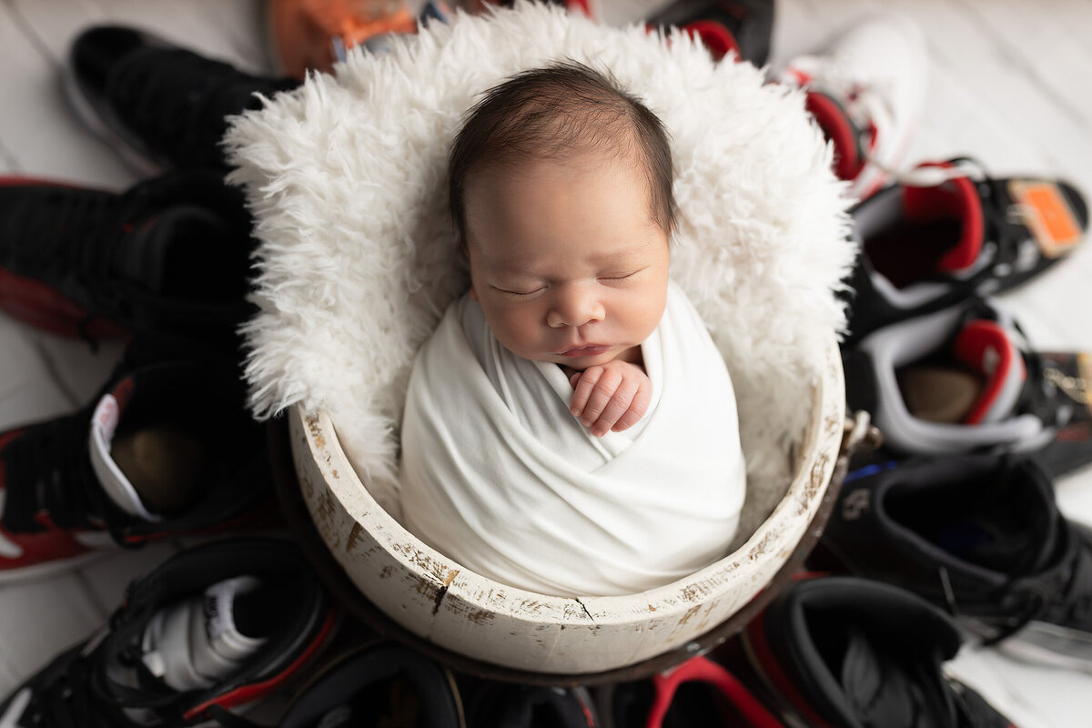 Close-up of a swaddled newborn posed in a white fluffy bowl, surrounded by high-end sneakers. DFW baby photographer offering unique newborn portraits.
