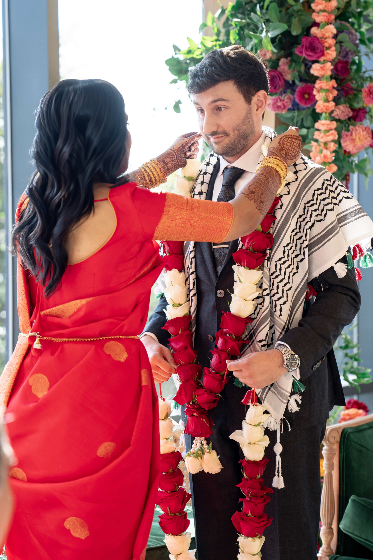A woman in a red sari places a garland around a mans neck during a ceremonial event. The man is wearing a suit and a patterned shawl. They are surrounded by colorful floral decorations.