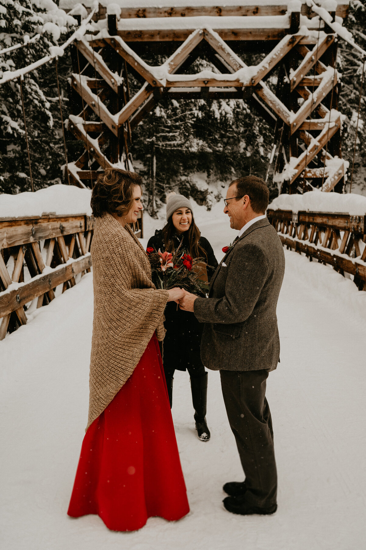Bride in red dress and groom holding hands during snowy elopement on historic bridge in Mount Rainier National Park!