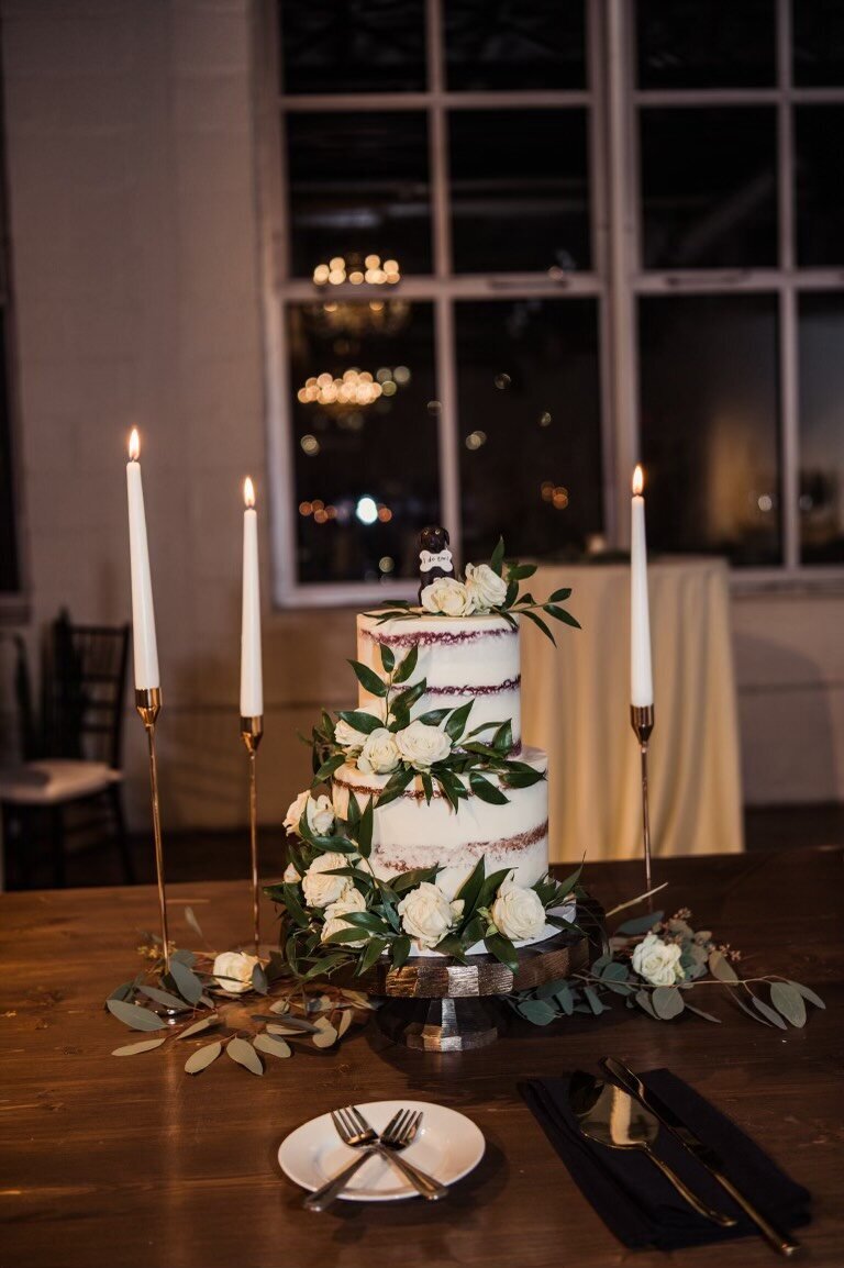 A two tier cake sits on a wooden table, surrounded by tapered candles and fresh greenery. The cake is naked style, with fresh greenery and white roses cascading down the left side and across the bottom. On top are two dog figurines, one black and one tan, wearing bandanas.