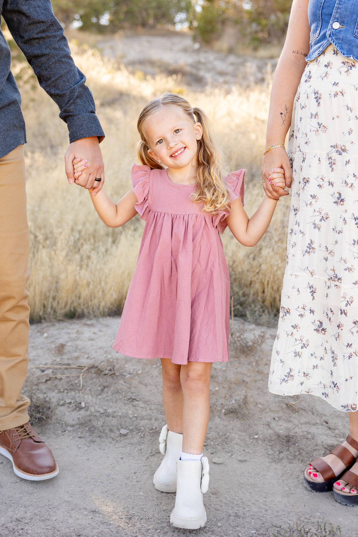 A young girl with blonde pigtails and a pink dress smiles brightly while holding both of her parents’ hands. The golden grass and desert scenery of Eagle Mountain, Utah, create a warm, rustic backdrop.