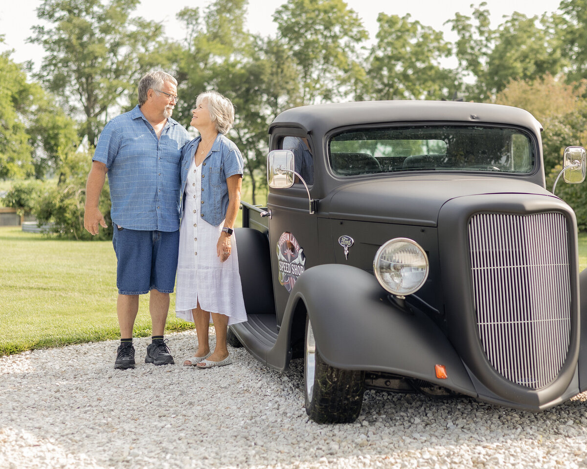Grandma and grandpa stadning next to a classic black car, standing next to each other in a rocked drive, smiling at each other.  Grandpa is wearing a light blue and white stripped short sleeve button up shirt, blue jean shorts, and has white short hair and goatee.  Grandma is wearing a white button up dress with a jean jacket over it, with short white hair.  