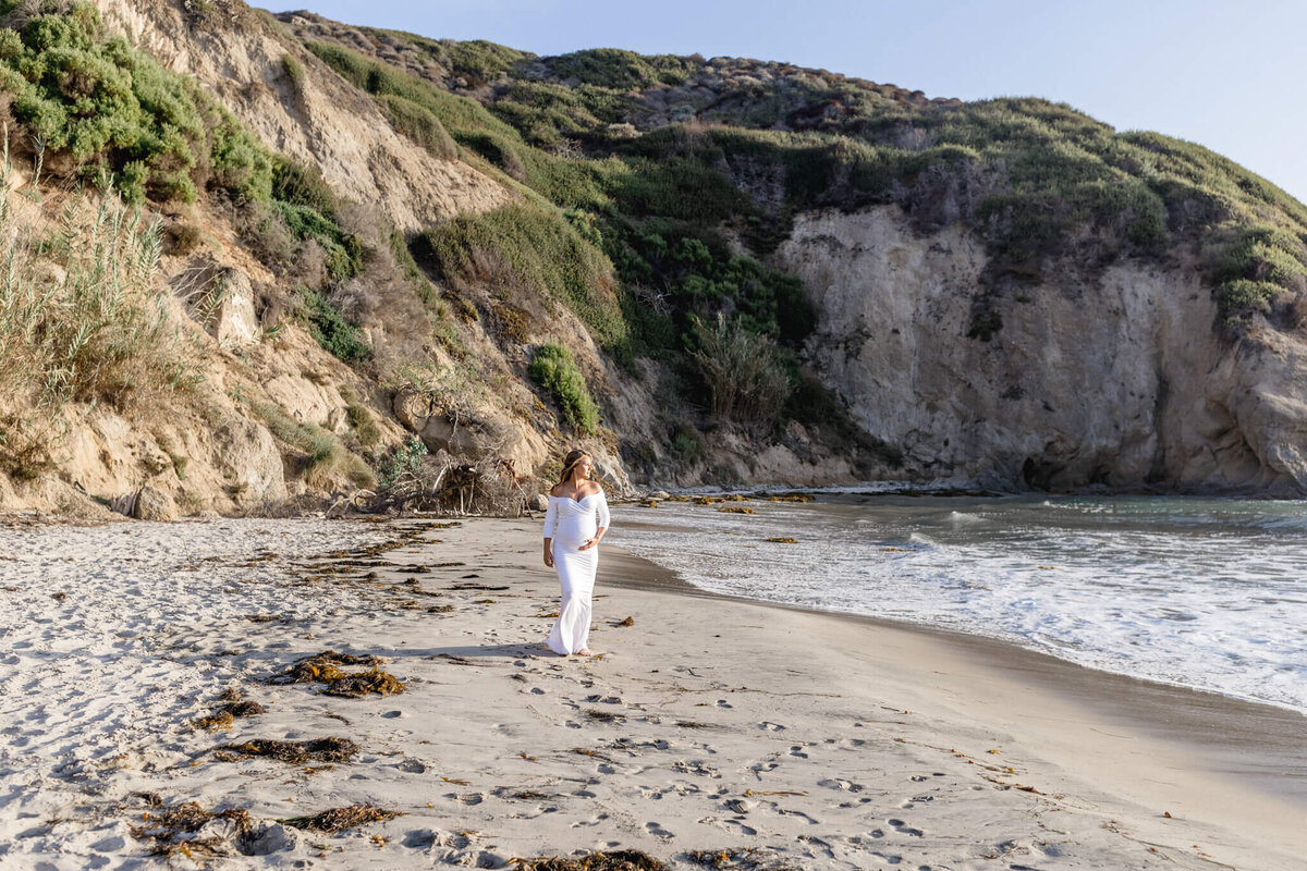 A person in white clothing walks along a sandy beach, with gentle waves lapping at the shore and rugged cliffs covered in greenery in the background. The sky is clear, suggesting a sunny day.