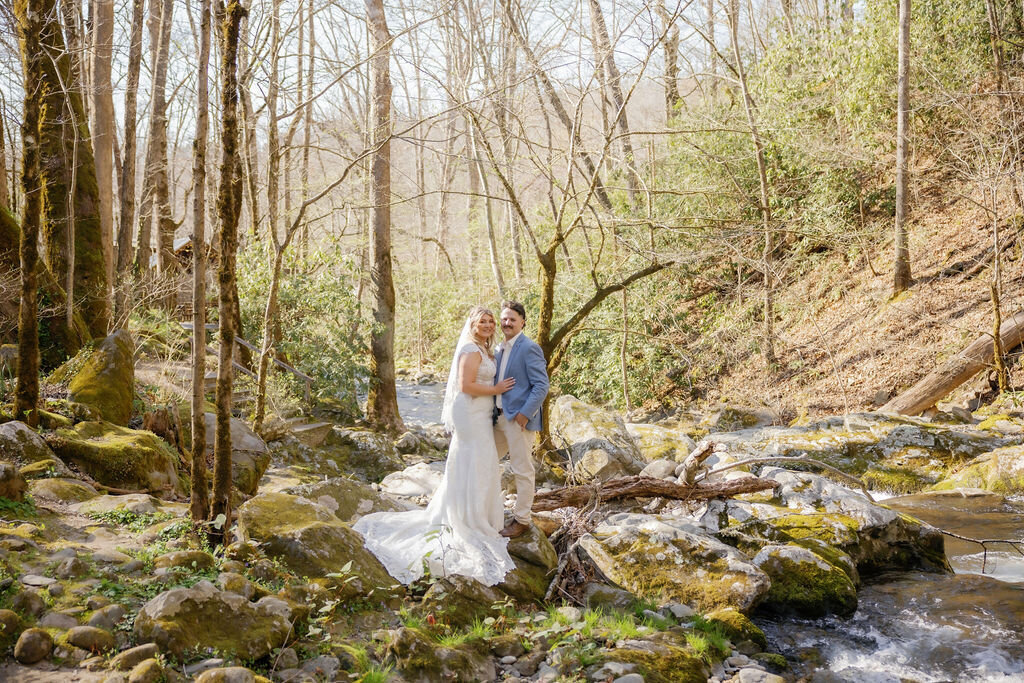 Gatlinburg elopement with bride and groom on the banks of a river in the woods at Ely's Mill with the morning sun shining through the trees and glistening the water