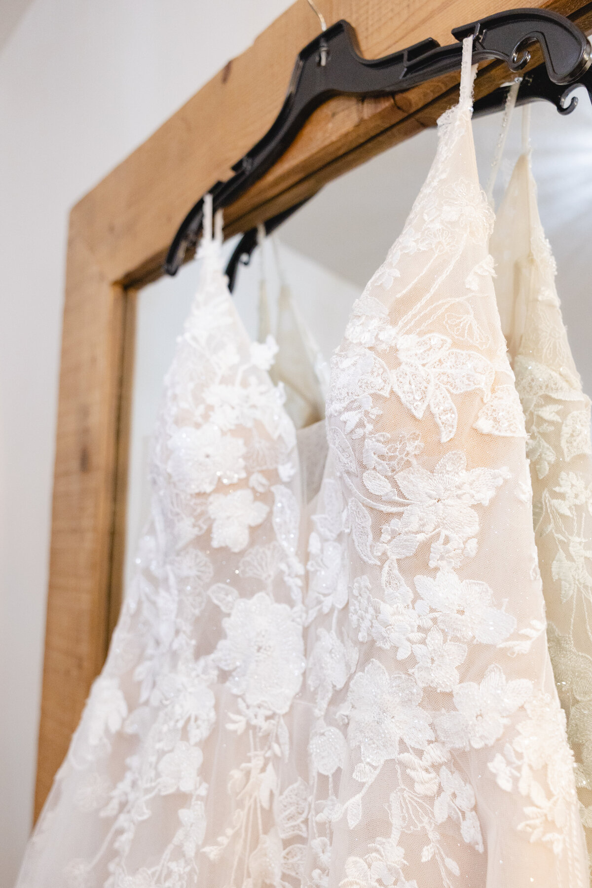 A beautifully detailed lace wedding dress hangs on a black hanger against a rustic wooden mirror, showcasing intricate embroidery and beading.